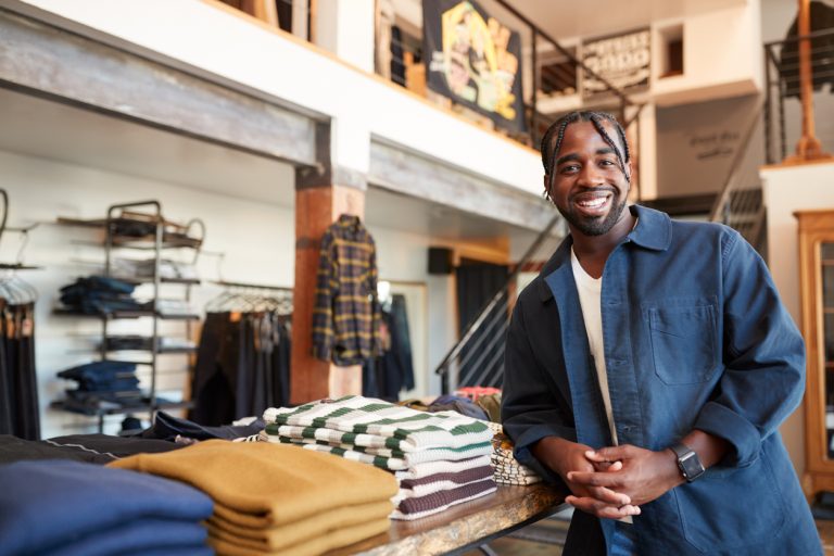 Portrait Of Smiling Male Owner Of Fashion Store Standing In Front Of Clothing Display
